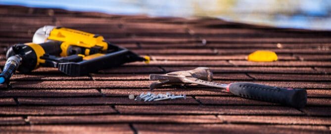 Hammer, nails, and a yellow nail gun resting on a shingle roof under sunlight.