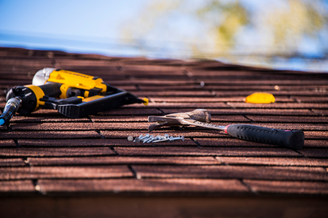 Hammer, nails, and a yellow nail gun resting on a shingle roof under sunlight.