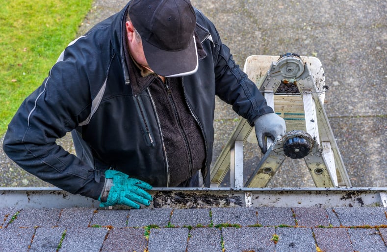 Man cleaning gutters while standing on latter and wearing gloves