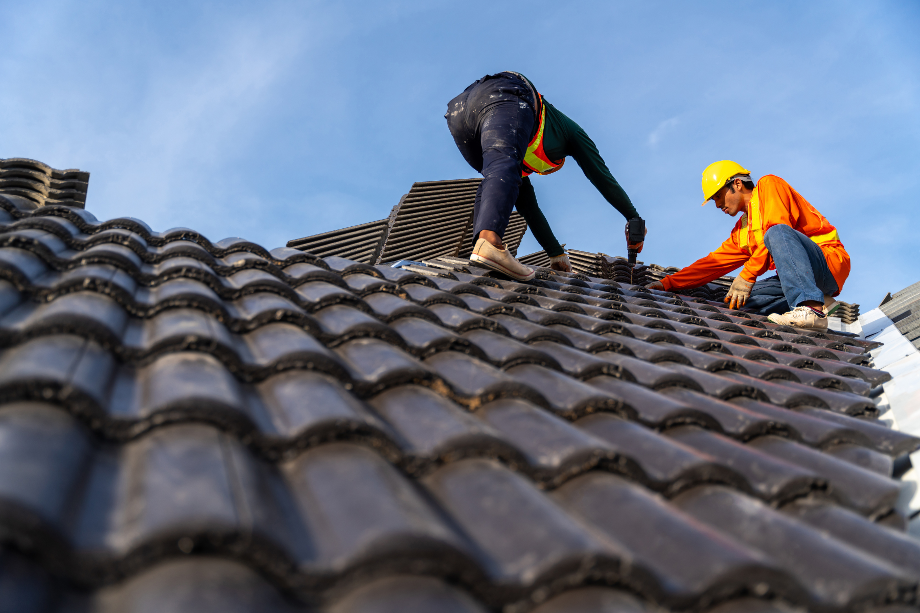 Two roofers working on a tiled roof under a clear blue sky, one in an orange safety jacket and hard hat, and the other installing tiles with a power tool.