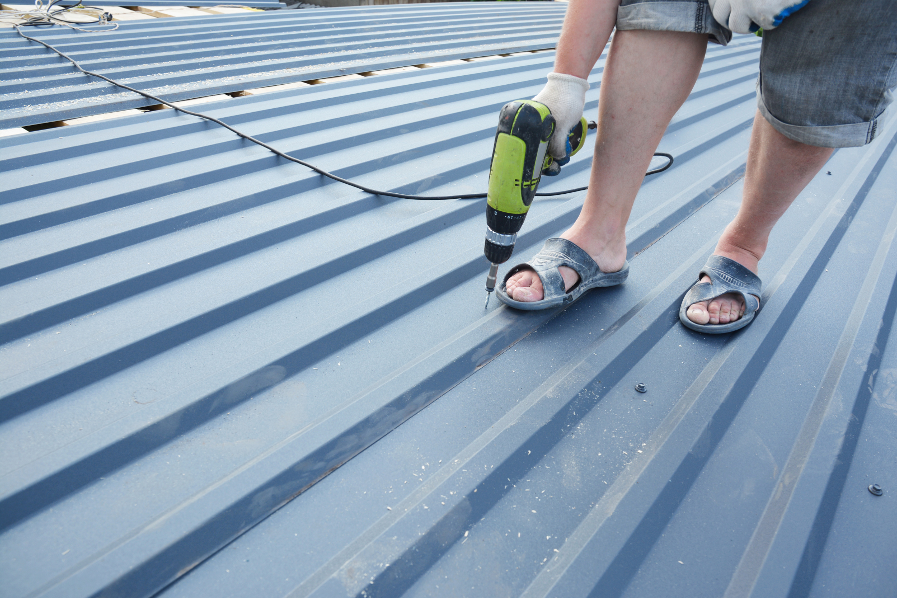 A person standing on a metal roof, holding a green electric drill. The person is wearing sandals and shorts, with visible tools and a power cord nearby.