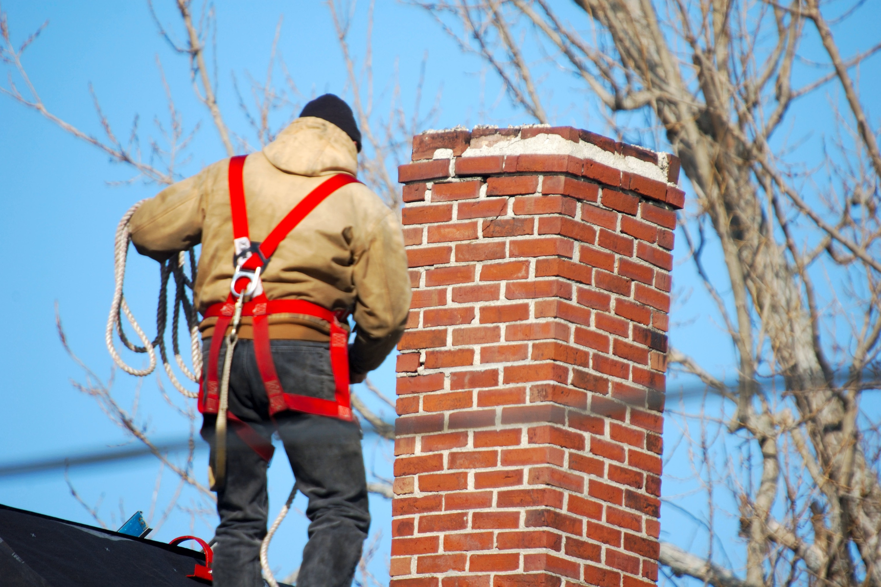 A construction worker wearing a safety harness and warm clothing standing on a roof beside a red brick chimney, preparing for chimney repairs. The background shows leafless tree branches against a clear blue sky.
