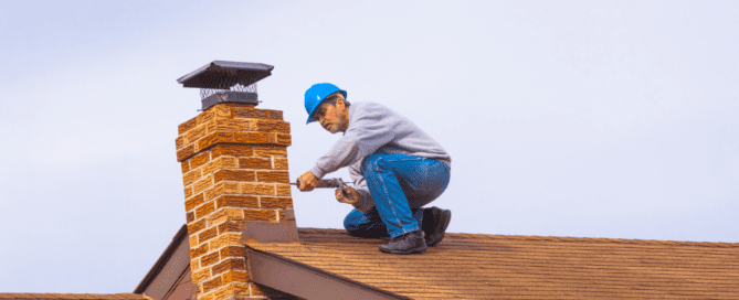 A construction worker wearing a blue hard hat and crouching on a sloped roof, performing repairs on a brick chimney. The worker is using tools to seal gaps in the chimney structure, with a chimney cap visible at the top. The roof is covered in brown shingles, and the sky in the background is overcast.