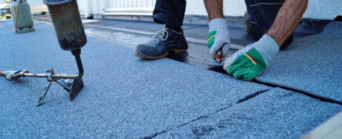 A person wearing gloves, jeans, and work boots working on a flat roof. They are using a tool to lay and seal a layer of roofing material. A propane torch and gas cylinder are visible in the background.