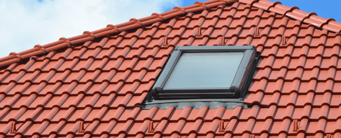 A red-tiled roof with a black framed skylight window installed near the center. The bright red tiles are neatly aligned, and the background shows a clear blue sky with a few white clouds.