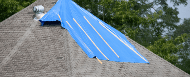 A house roof with a large blue plastic tarp secured over part of the shingles. The tarp is fastened with wooden planks, covering a damaged area. Trees are visible in the background.