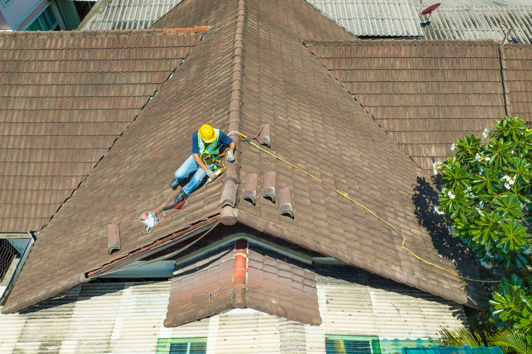 A person wearing a safety harness, yellow helmet, and high-visibility vest repairing a tiled roof. They are seated at the roof's peak, surrounded by tools and safety ropes.