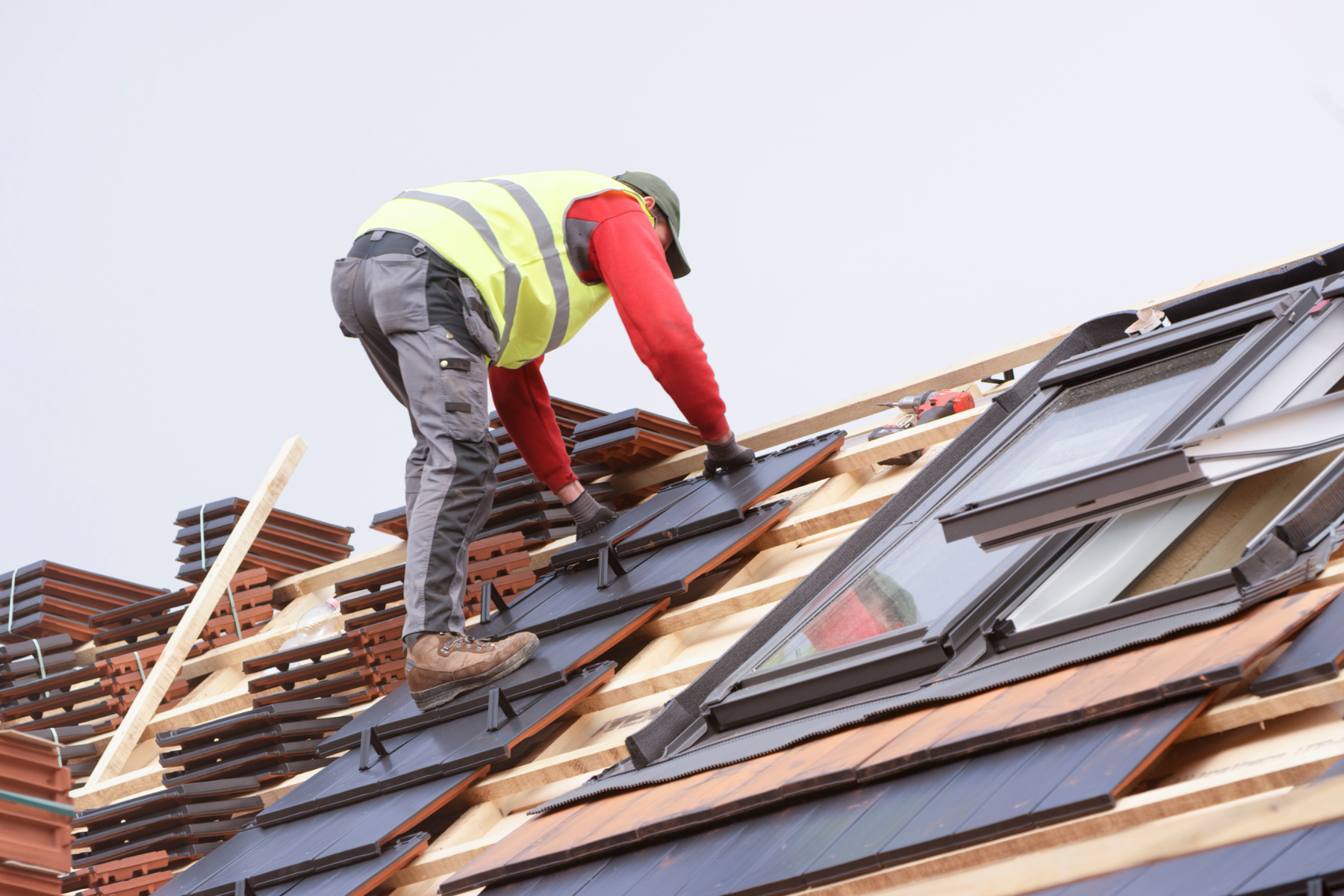  A construction worker wearing a high-visibility vest and protective gear is installing solar panels on a sloped rooftop. The roof frame is partially covered with black solar tiles, and an open skylight window is visible on the right side.
