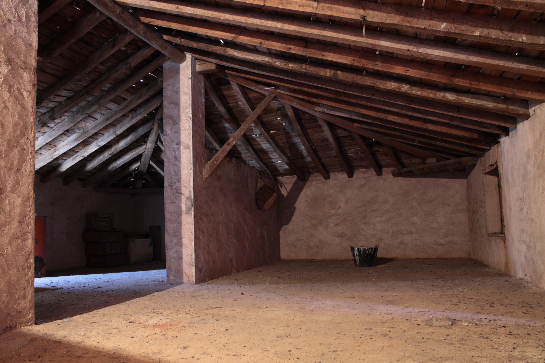 A rustic attic with wooden beams, rough plastered walls, and a dirt floor. A basket and crates are stored in the space, with natural light streaming through openings in the structure.