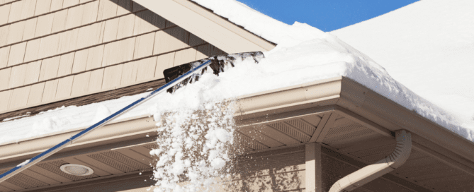 A beige house roof with snow being cleared using a long-handled snow rake. Snow is falling off the roof onto the ground. The roof has beige siding and a white gutter system.
