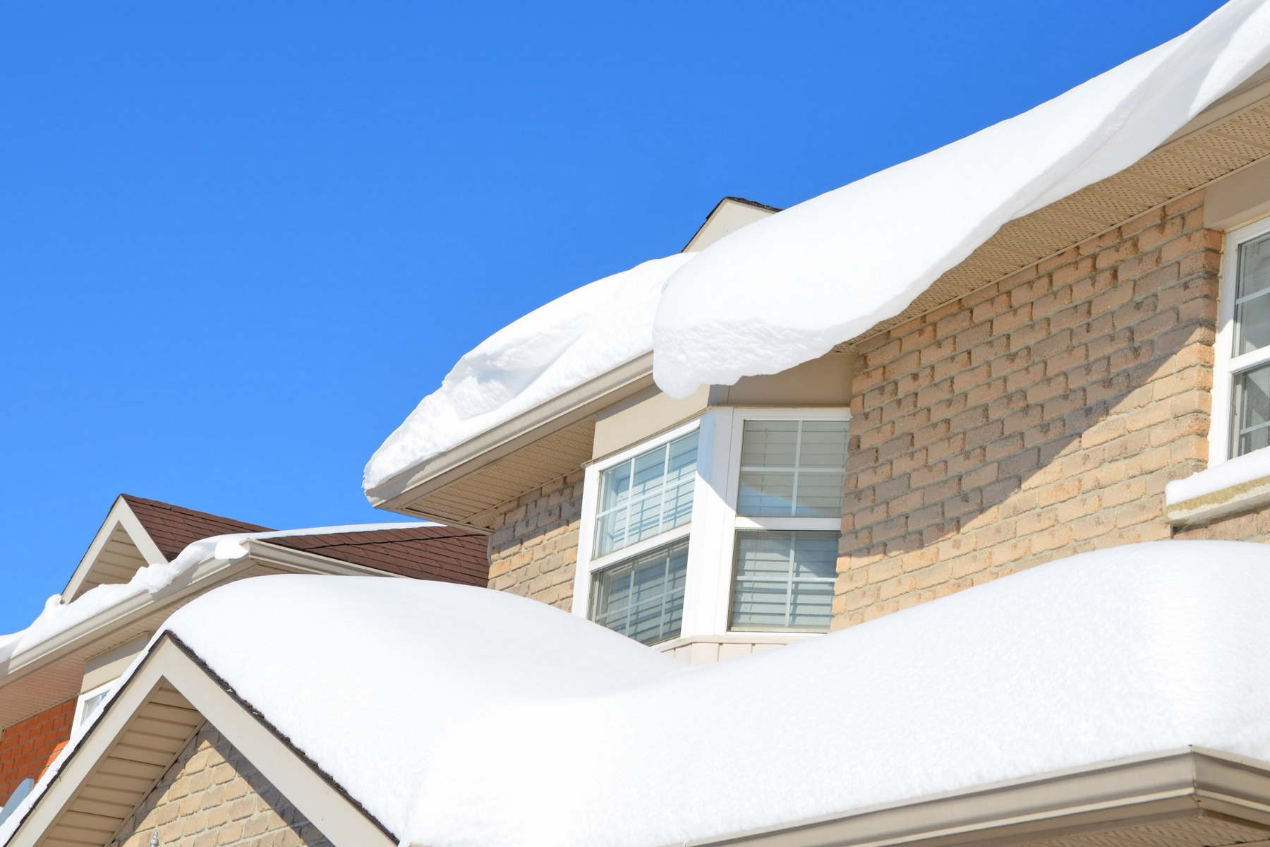 A close-up of a house roof covered in thick, heavy snow. The house is made of light-colored bricks, and the windows have white shutters. The sky is bright blue with no clouds.