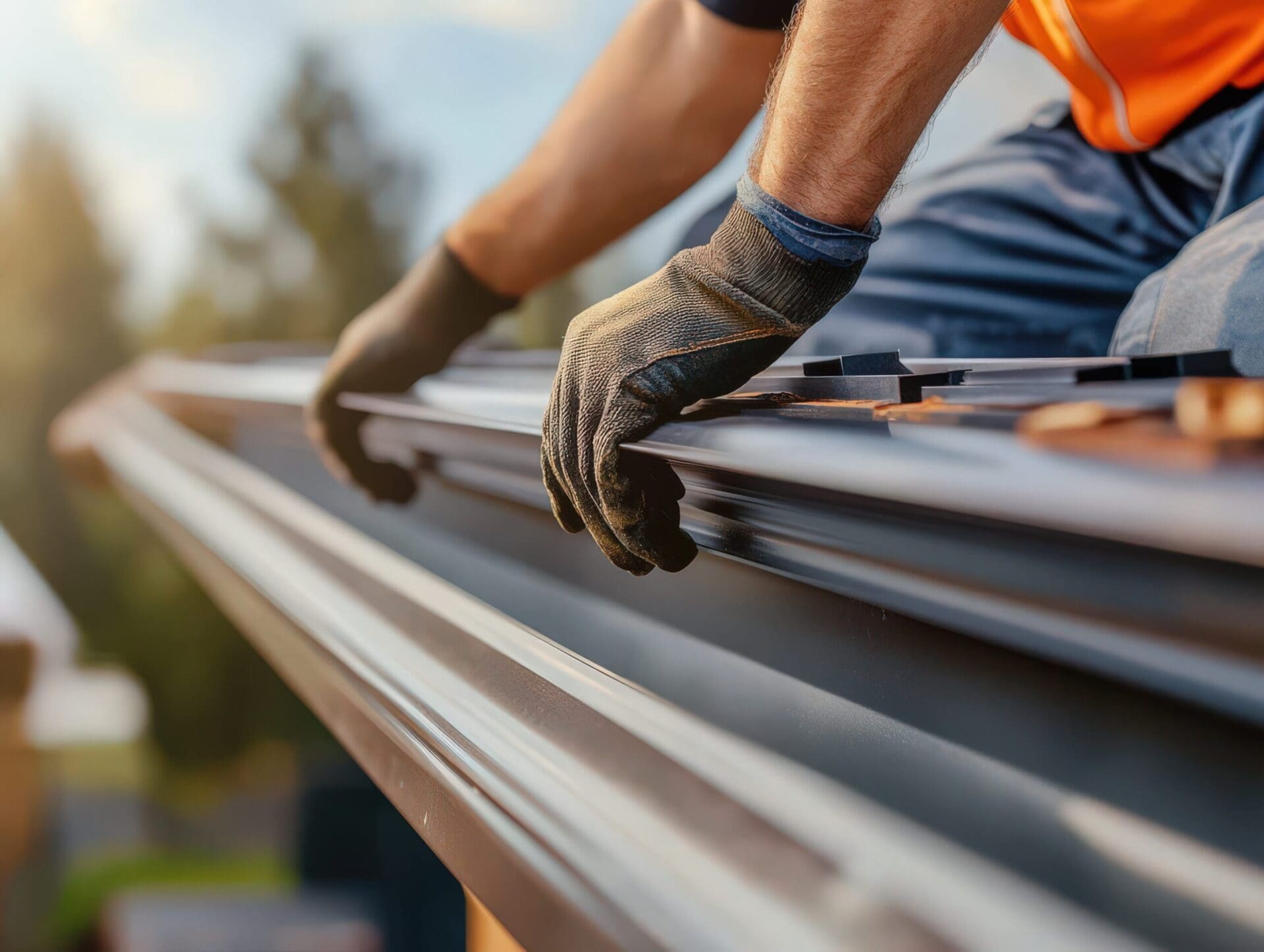 A close-up of a worker's gloved hands installing a metal gutter system along the edge of a roof.