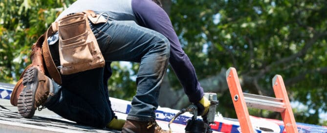 A worker wearing protective gear is installing shingles on a roof using a nail gun.