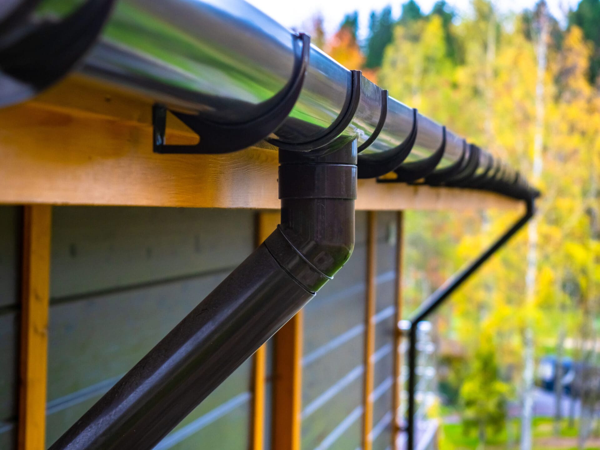 A shiny metal gutter with a downspout system installed on a wooden building, surrounded by autumn trees in the background.