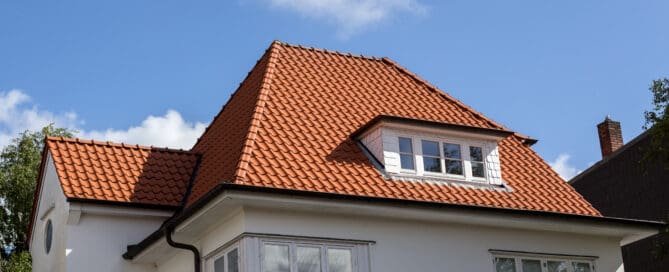 A house with a distinct red clay tile roof, featuring multiple peaks and a dormer window. The roof tiles are neatly arranged, and the structure has a classic architectural design with white walls and dark gutters.