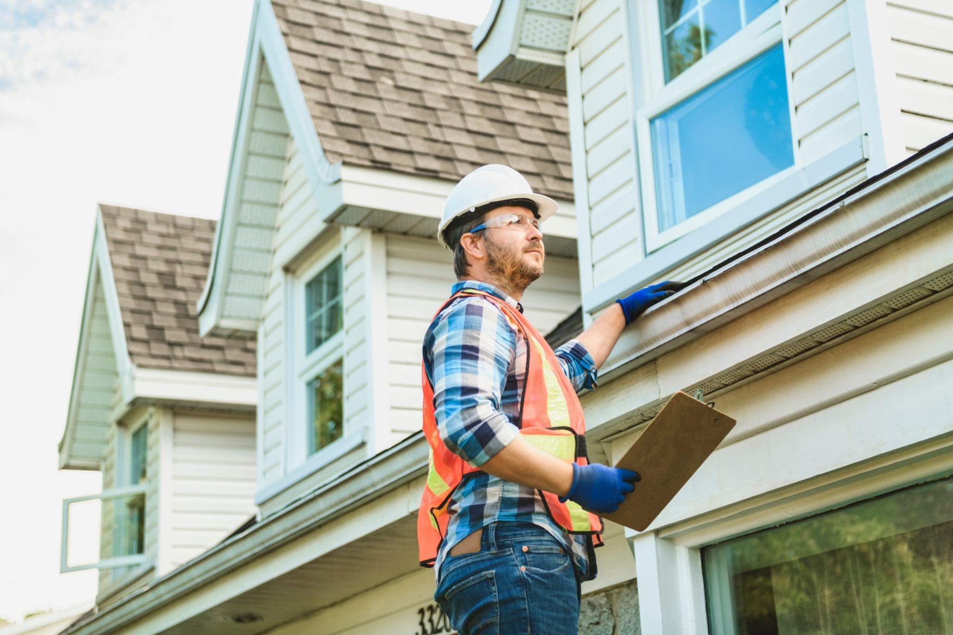 A man with hard hat standing on steps inspecting house roof.