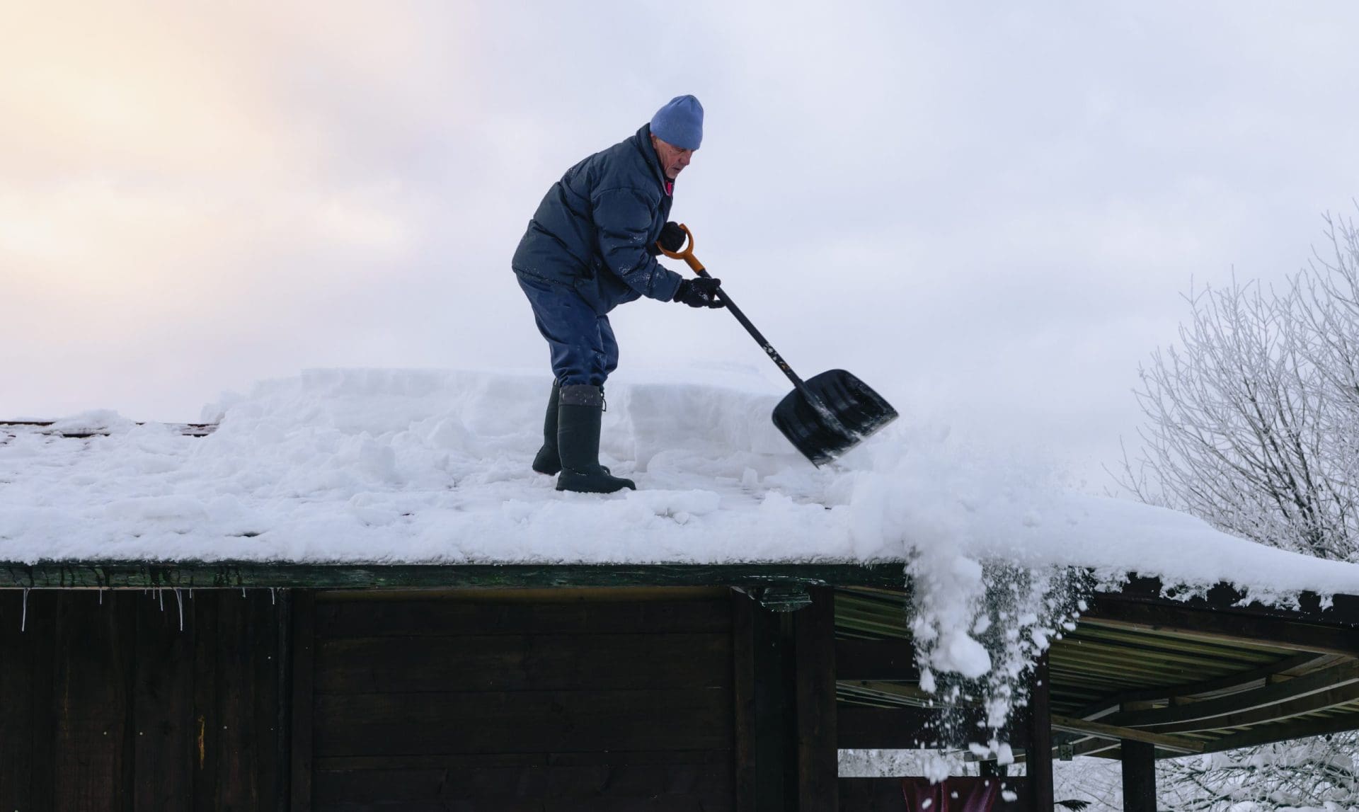 A person in winter clothing and boots is shoveling thick snow off a dark roof. The snow is being thrown over the edge, with a cloudy winter sky in the background.