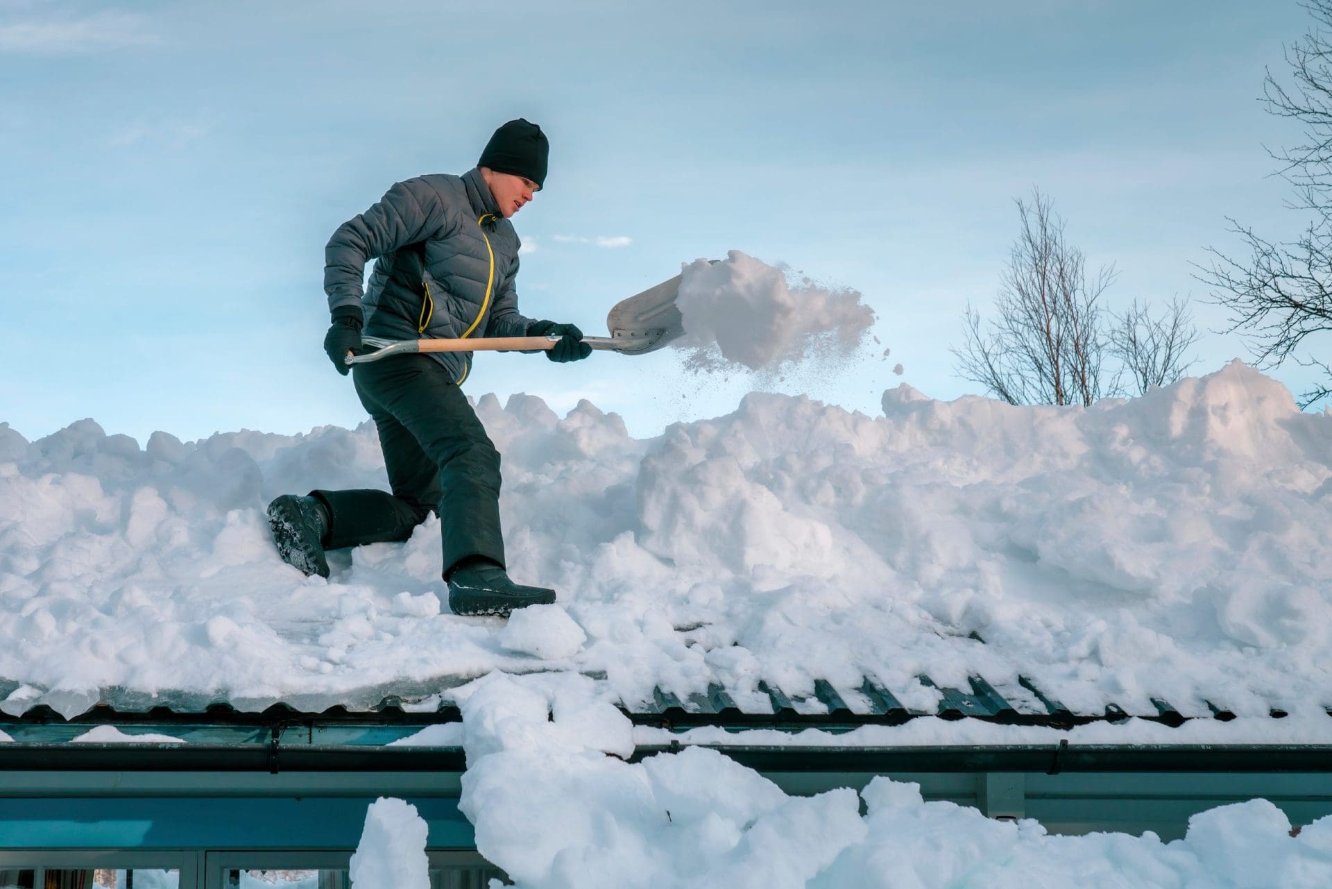 Person with shovel remove snow from roof of totally snow covered house, cabin in mountains. Cloudy warm day, much fresh snow after snowstorm