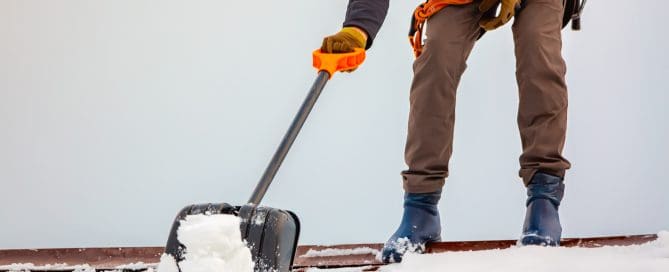 A person wearing gloves and winter boots is clearing snow from a rooftop using a snow shovel with an orange handle. The individual is secured with safety gear, and snow is visibly being pushed off the roof.