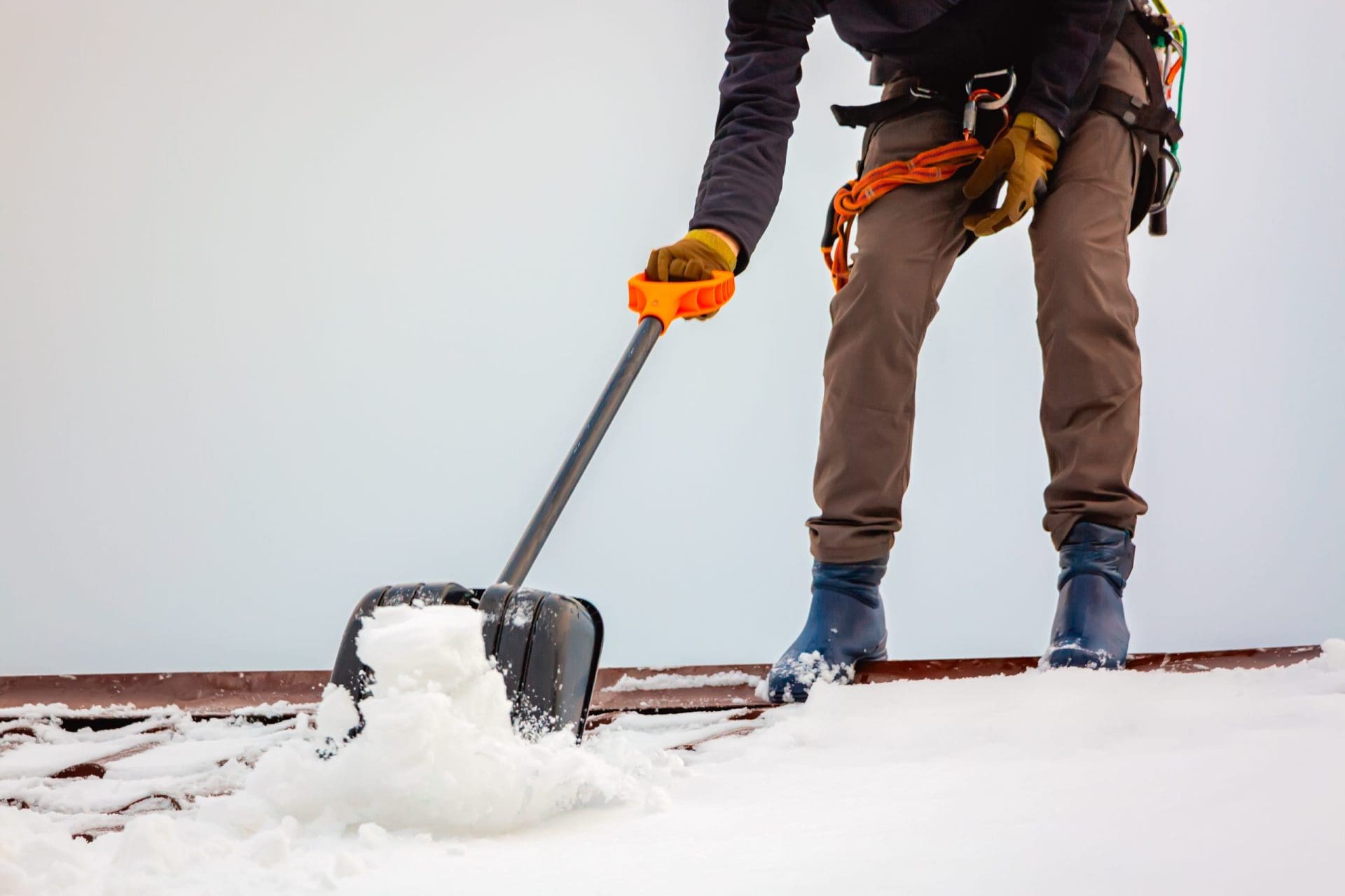 A person wearing gloves and winter boots is clearing snow from a rooftop using a snow shovel with an orange handle. The individual is secured with safety gear, and snow is visibly being pushed off the roof.