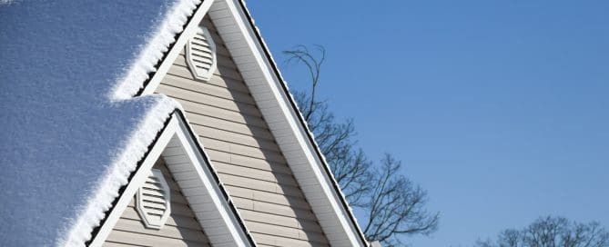 Steep gable rooftops coated in snow on a bright winter day, with trees in the background under a clear sky.