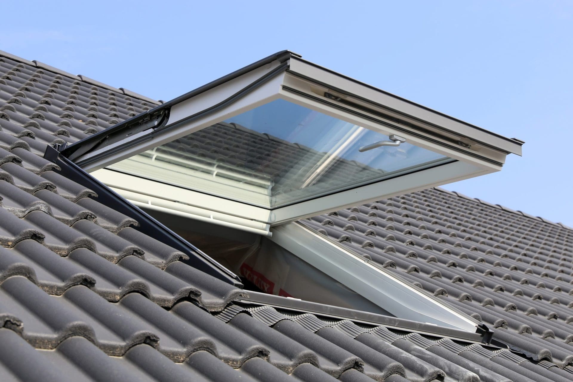 A modern skylight window installed on a tiled roof, tilted open to allow ventilation and sunlight.