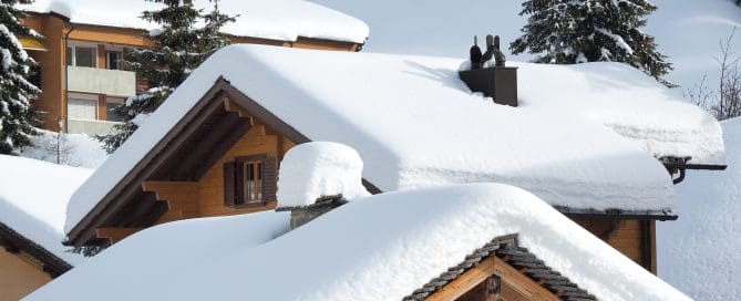Wooden chalet-style homes with steep roofs blanketed in thick, heavy snow, surrounded by snow-covered trees.
