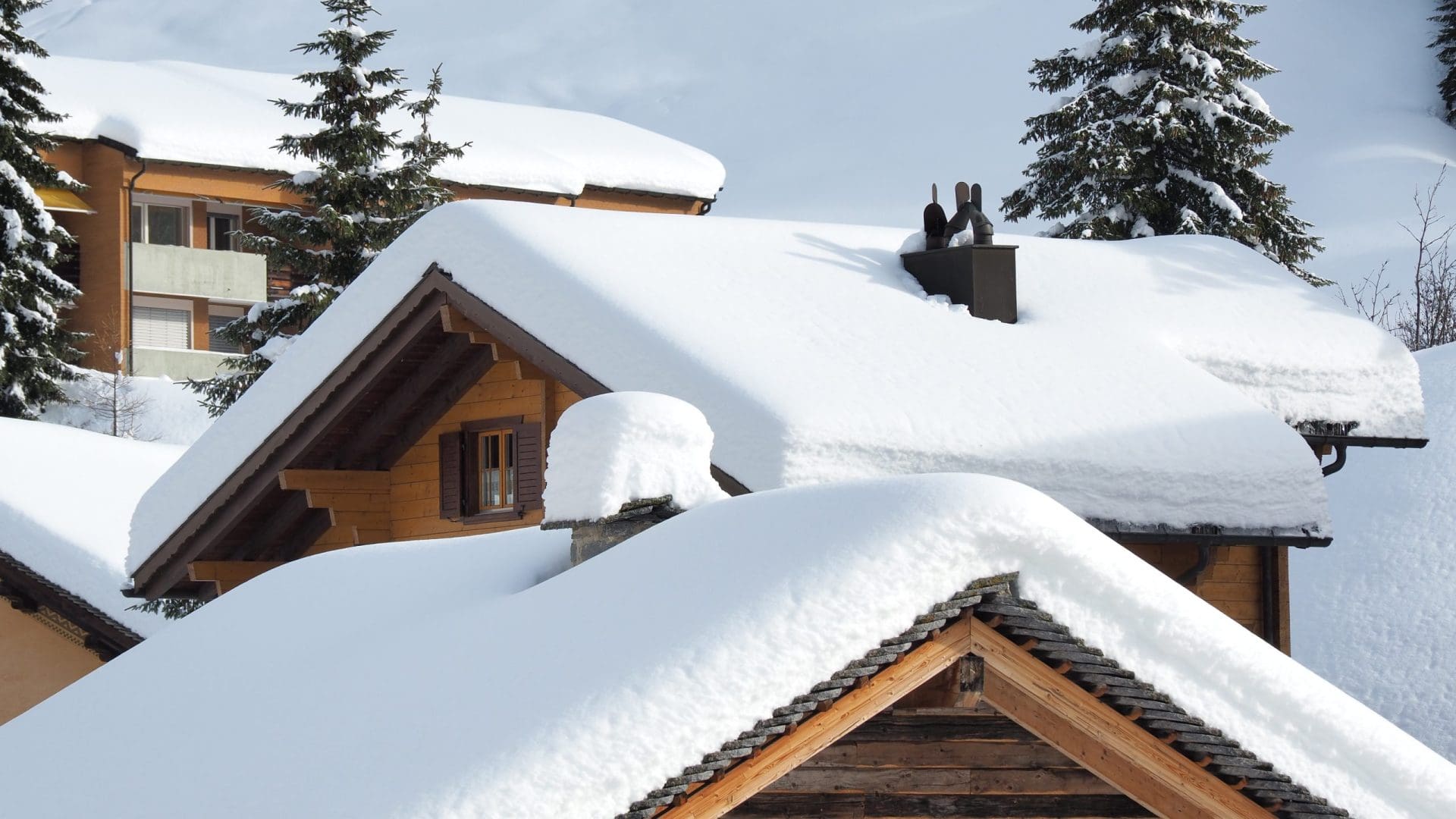 Wooden chalet-style homes with steep roofs blanketed in thick, heavy snow, surrounded by snow-covered trees.