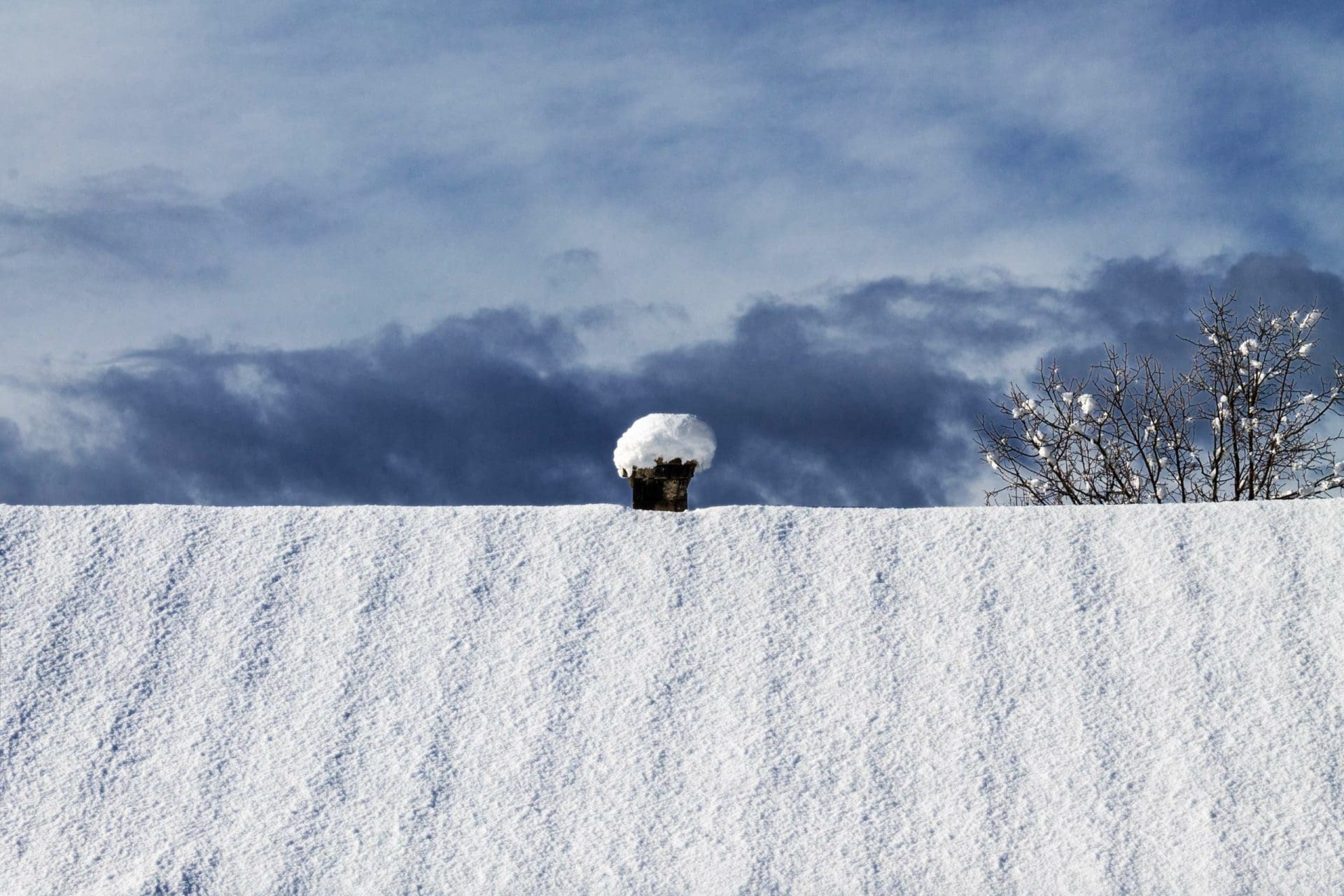 A rooftop fully covered in fresh snow with a chimney capped by a rounded pile of snow, set against a dramatic cloudy sky.
