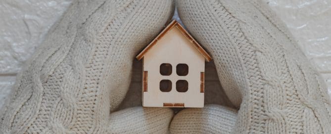 House insulation concept with hands wearing woolen mittens holding a wooden model of house.