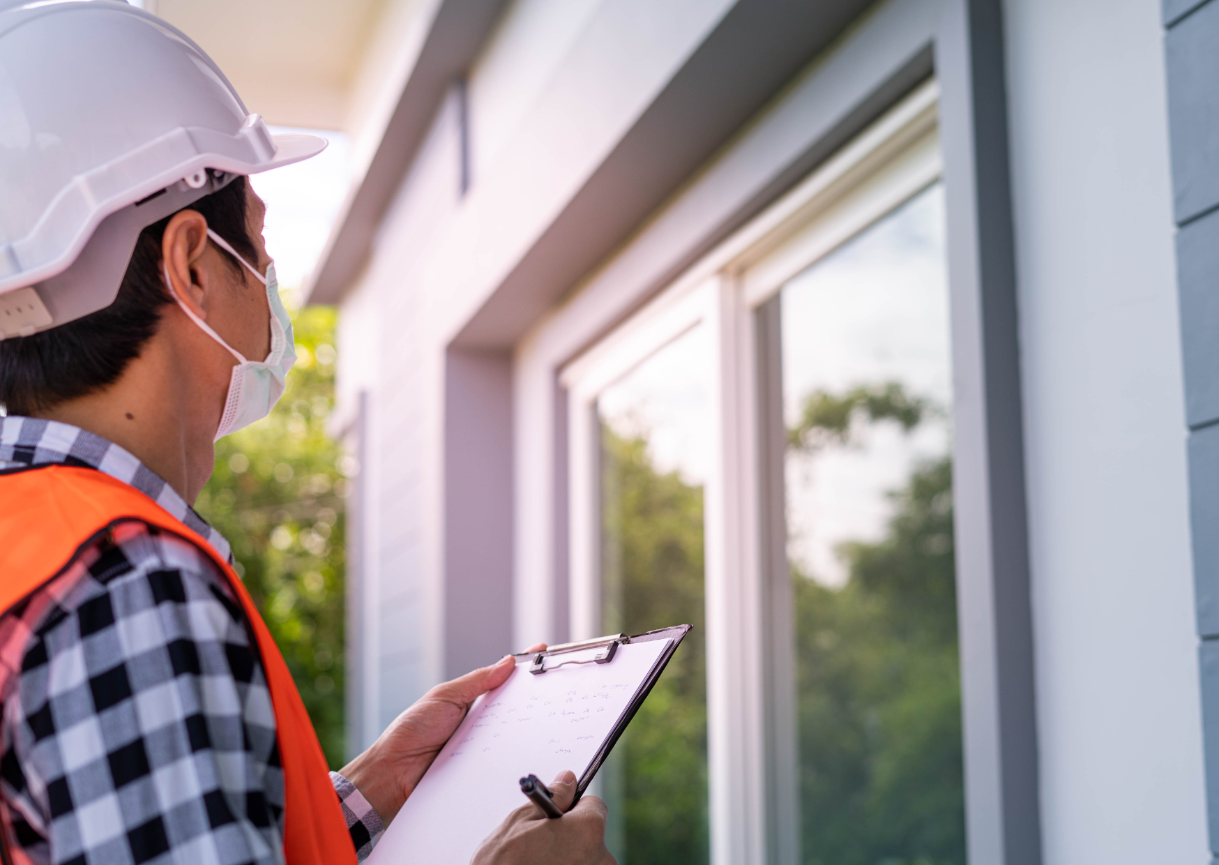 A person wearing a helmet and mask holding a clipboard while looking at a building exterior.