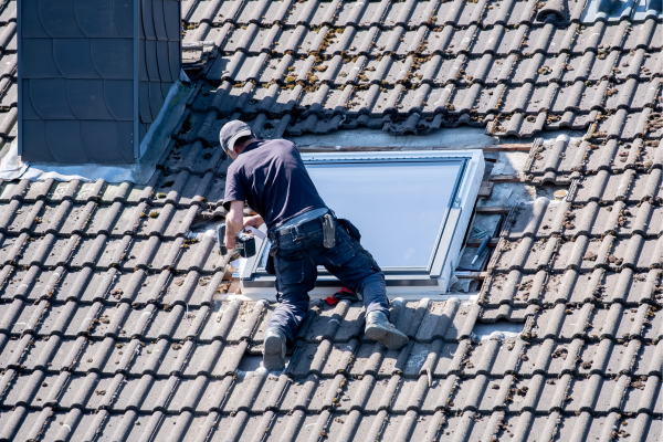  worker in a dark shirt, cap, and tool belt kneels on a weathered tile roof, using a cordless drill to install or repair a skylight unit. Surrounding tiles are removed around the opening, exposing the wood battens beneath. A dark dormer or chimney sits in the upper left.
