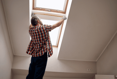 Inside a room with a sloped white ceiling, a man in a red plaid shirt and jeans reaches up with both hands to open or adjust a skylight window. The angle is from below, looking up toward the skylight.