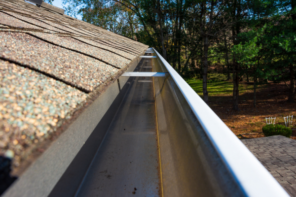 Close-up view down the length of a clean metal gutter running along the edge of an asphalt shingle roof. The gutter is empty and debris-free, with bare trees and a wooded yard in the background under daylight.
