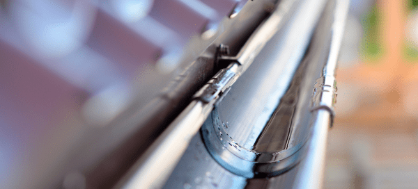 Tight macro shot of a metal rain gutter and its bracket, with sunlight reflecting off the curved surface. The roof shingles and background are heavily blurred, drawing focus to the gutter's edge and seam.
