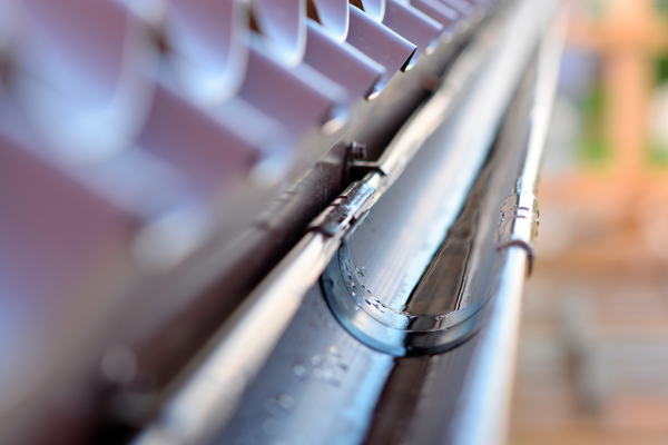 Tight macro shot of a metal rain gutter and its bracket, with sunlight reflecting off the curved surface. The roof shingles and background are heavily blurred, drawing focus to the gutter's edge and seam.