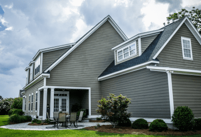 Exterior view of a large gray house with horizontal lap siding, white trim, and dark shingle roofing across multiple gables and dormers. A covered patio with a table and chairs sits at the rear, bordered by a paved walkway, shrubs, and a green lawn under a partly cloudy sky.