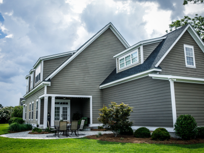 Exterior view of a large gray house with horizontal lap siding, white trim, and dark shingle roofing across multiple gables and dormers. A covered patio with a table and chairs sits at the rear, bordered by a paved walkway, shrubs, and a green lawn under a partly cloudy sky.