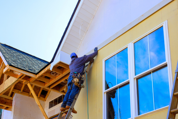 A worker in a blue shirt, jeans, hard hat, and tool belt stands on a ladder, fitting trim or siding under the eave of a house. The wall shows pale yellow sheathing, and a large window beside him reflects blue sky and clouds. Wood framing and dark shingles are visible to the left.