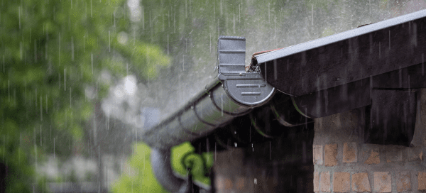 Rain pours down onto a metal gutter and downspout attached to a brick home. Green trees are visible in the blurred background, and water runs along the edge of the roofline.