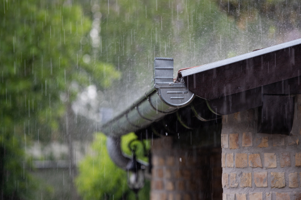 Rain pours down onto a metal gutter and downspout attached to a brick home. Green trees are visible in the blurred background, and water runs along the edge of the roofline.