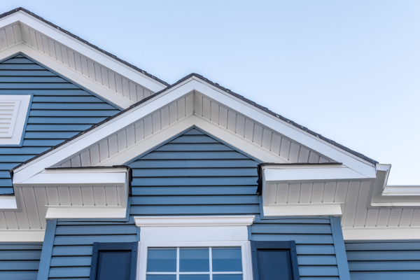 Close-up of a home's upper exterior showing two gabled peaks. The walls combine horizontal blue lap siding with vertical board-and-batten siding in the gable points, accented by white trim. A louvered vent and a shuttered window are partly visible against a clear sky.