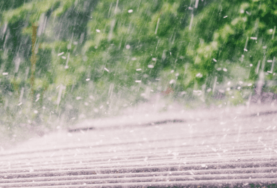 Close-up of rain falling hard onto a ridged roof surface, with droplets splashing on impact. Green foliage is blurred in the background, conveying an active storm.
