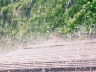 Close-up of rain falling hard onto a ridged roof surface, with droplets splashing on impact. Green foliage is blurred in the background, conveying an active storm.
