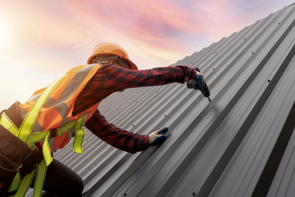 A worker wearing a hard hat, reflective safety vest, harness, and gloves uses a power drill to secure a screw into a gray metal roofing panel. The sun sets behind them, casting an orange and pink glow across the sky.
