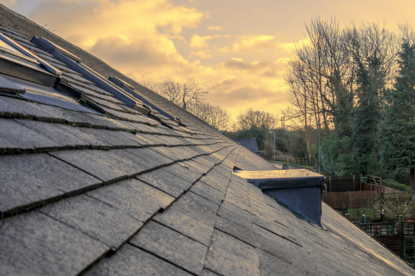A close-up view along the ridge of an aged tile roof, showing textured tiles with some visible wear and metal flashing. Bare trees and a fenced yard are visible in the background under a golden sunset sky.