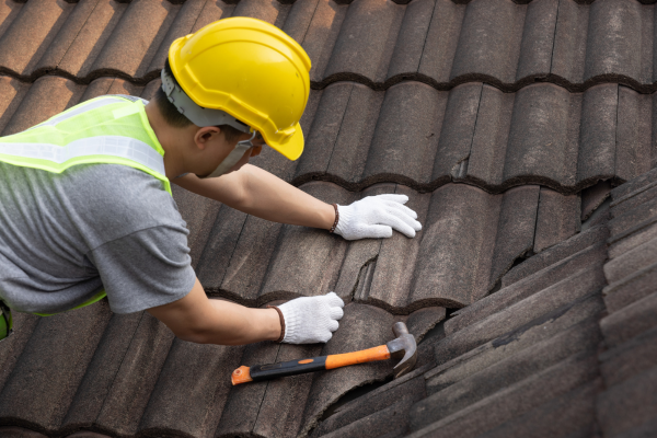 A worker wearing a yellow hard hat, safety glasses, reflective vest, and white gloves kneels on a brown tile roof and holds a hammer while working on a damaged section of tile.
Image 3
Title: Modern White Home Exterior at Sunset
Description: A modern two-story white home with large windows, a covered patio, and a small second-floor balcony. The setting sun shines through trees in the background, and a landscaped lawn with flowering plants is visible in the foreground.