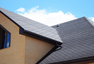 A close-up view of a home's dark asphalt shingle roof with multiple gables, gutters, and a small roof vent. The exterior features tan brick siding, and a partly cloudy blue sky is visible in the background.