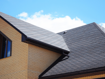 A close-up view of a home's dark asphalt shingle roof with multiple gables, gutters, and a small roof vent. The exterior features tan brick siding, and a partly cloudy blue sky is visible in the background.