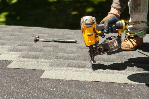 A close-up of a person wearing gloves and work boots using a yellow pneumatic nail gun to secure gray asphalt shingles to a rooftop. A hammer rests on the shingles nearby, and trees are visible in the background.